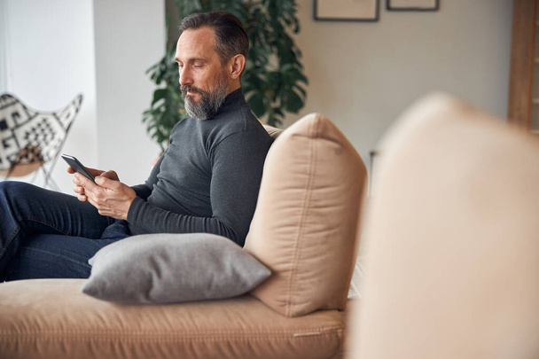 Caucasian adult man is sitting with smartpone in modern cozy office lounge zone - 写真・画像