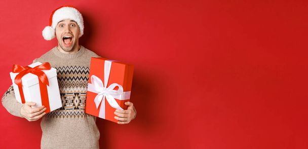Image of cheerful attractive guy holding christmas presents, standing in santa hat and winter sweater, smiling amazed, standing over red background - Foto, Bild