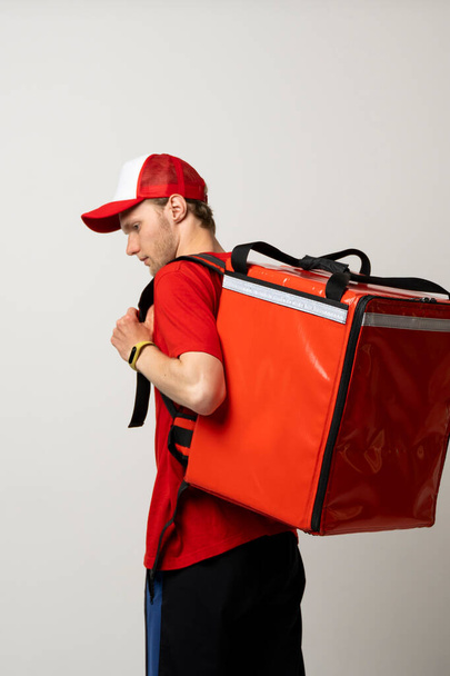Служба доставки быстрого питания Cocnept. Портрет улыбающегося курьера в красной униформе с красным терморюкзаком на фоне белой студии - Фото, изображение
