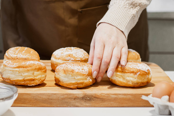 Woman prepares fresh donuts with jam in home kitchen. Cooking traditional Jewish Hanukkah sufganiyot. Hands sprinkle Berliners with powdered sugar. - Фото, изображение