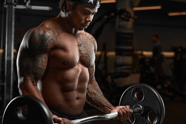 Сильный и мускулистый темнокожий мужчина тренируется на современном оборудовании в тренажерном зале. Портрет накачанного тренера по фитнесу - Фото, изображение
