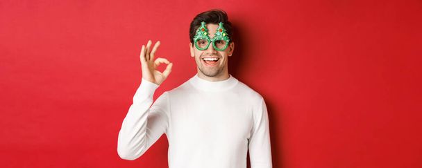 Feliz chico caucásico deseando feliz Navidad, celebrando las vacaciones de invierno, mostrando signo de bien y sonriendo complacido, de pie sobre el fondo rojo - Foto, imagen