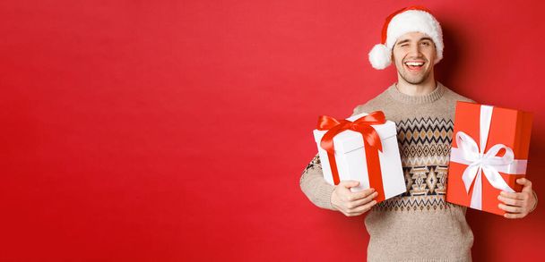 Έννοια των χειμερινών διακοπών, το νέο έτος και γιορτή. Πορτρέτο του αυτοπεποίθηση και αναιδής νεαρός άνδρας ετοιμάσει δώρα για τα Χριστούγεννα, κλείσιμο του ματιού και κρατώντας δώρα, στέκεται πάνω από το κόκκινο φόντο - Φωτογραφία, εικόνα