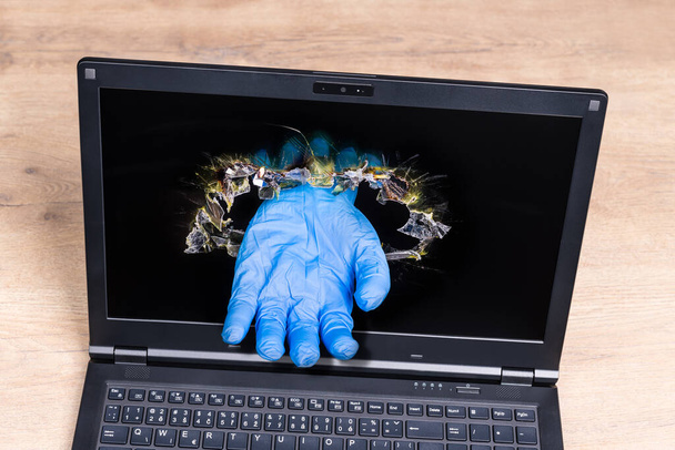 Laptop-Detail mit imaginärer helfender Hand in blauem Handschuh, der aus einem Loch im Display ragt. Medizinische Online-Hilfe. Symbolischer virtueller Arzt. Fernbehandlung bei Coronavirus-Erkrankungen. - Foto, Bild