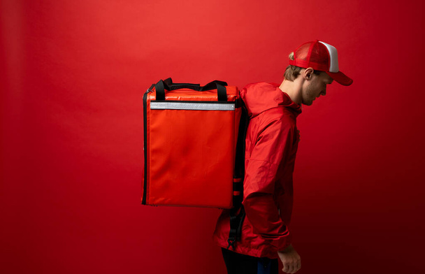 Side view of young courier in a red uniform and with a thermo bag on color red background, space for text. Food delivery service. - Foto, Imagen