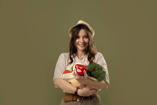Portrait of happy smiling young woman in beige oversize t-shirt holding reusable string bag with groceries over green background. Sustainability, eco living and people concept. - Foto, Imagen