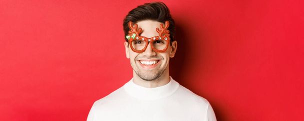 Konzept der Winterferien, Weihnachten und Feiern. Nahaufnahme eines attraktiven brünetten Kerls mit Parteibrille, der vor rotem Hintergrund lächelt und glücklich aussieht - Foto, Bild