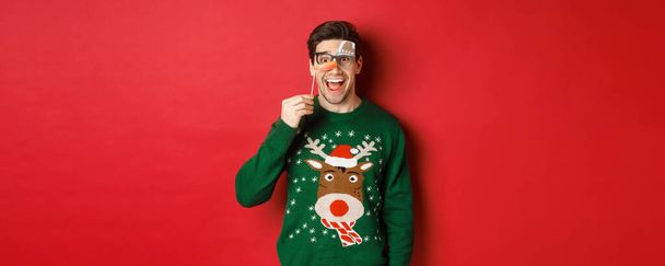 Porträt eines amüsierten gutaussehenden Mannes im Weihnachtspulli, der eine lustige Party-Maske hält und lächelt, Winterurlaub feiert, vor rotem Hintergrund steht - Foto, Bild