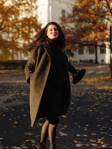 Jonge mooie zwangere vrouw met donker haar in een zwarte strakke jurk en jas poseren op een herfstweide in het park - Foto, afbeelding