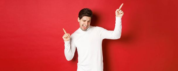 Porträt eines gutaussehenden Mannes, der Silvesterparty genießt, tanzt und Spaß hat, mit erhobenem Zeigefinger über rotem Hintergrund steht - Foto, Bild