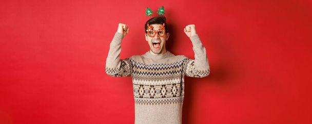 Portrait d'homme ravi et heureux dans des verres de fête, pull d'hiver, criant de joie et levant les mains vers le haut, remportant le prix de Noël, atteindre un nouvel objectif de l'année, debout fond rouge - Photo, image