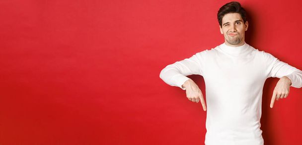 Портрет привлекательного, скептически настроенного молодого человека в белом свитере, гримасничающего и указывающего пальцем вниз, не согласного и не любящего что-то, стоящего на красном фоне - Фото, изображение