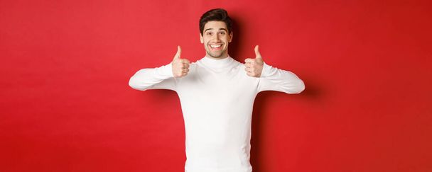 Alegre modelo masculino guapo en suéter blanco, mostrando pulgares hacia arriba en la aprobación, como algo bueno, de pie sobre el fondo rojo y sonriendo complacido - Foto, Imagen