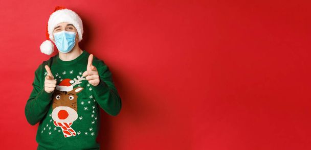Koncepcja nowego roku, covid-19 i dystans społeczny. Wesoły mężczyzna w świątecznym swetrze, masce medycznej i czapce Mikołaja, wytykający palcami kamerę, życzący szczęśliwych świąt, stojący nad czerwonym tłem - Zdjęcie, obraz