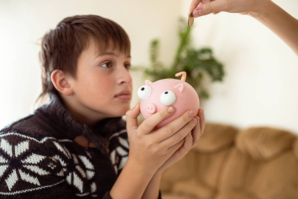 Junge mit Sparschwein bettelt um Geld. Hand wirft eine Münze in ein Sparschwein. Teenager fordern Geld, um Geld zu sparen. Persönliche Ausgaben von Schülern. - Foto, Bild