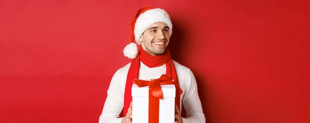 Concepto de vacaciones de invierno, Navidad y estilo de vida. Primer plano de hombre atractivo en sombrero de santa y bufanda, celebración de regalo de año nuevo, mirando a la derecha y sonriendo, de pie sobre el fondo rojo - Foto, imagen