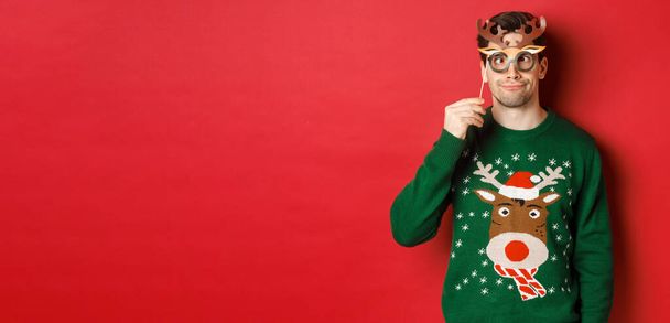 Zabawny człowiek w świątecznym swetrze i masce imprezowej, świętujący ferie zimowe, pokazujący śmieszne twarze, stojący na czerwonym tle - Zdjęcie, obraz