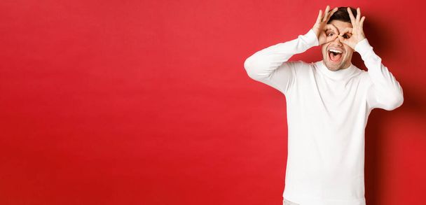 Portrait of handsome guy in white sweater, making funny mask with fingers, looking happy and smiling, standing over red background - Foto, Imagen