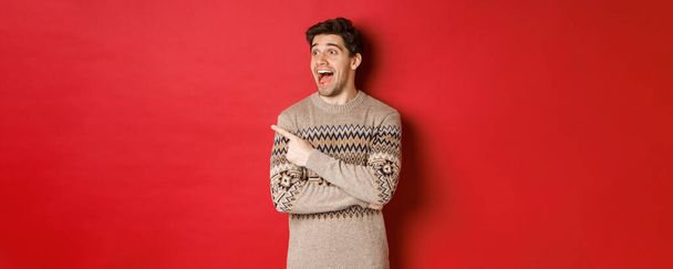 Portrait of attractive caucasian man celebrating new year, winter holidays, pointing finger and looking left with amazed expression, showing christmas advertisement, standing over red background - Foto, Imagen