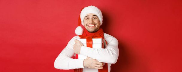 Konzept für Winterurlaub, Weihnachten und Lifestyle. Bild von schönen Kerl mit Weihnachtsmütze und Schal, umarmt sein Neujahrsgeschenk und lächelt geschmeichelt, steht über rotem Hintergrund - Foto, Bild