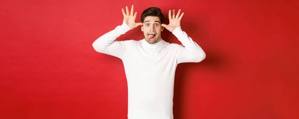 Portrait of funny caucasian guy, showing tongue and making faces, wearing white sweater, standing against red background - Фото, изображение