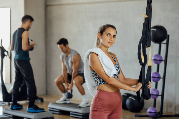 Confident Fitness woman standing in gym with two men exercising in the back. - Photo, Image