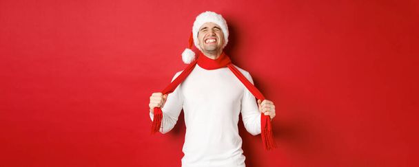 Noel Baba şapkalı sıkıntılı ve hayal kırıklığına uğramış bir adamın resmi. Üzüntü eşarbıyla kendini boğuyor, kırmızı arka planda duruyor. - Fotoğraf, Görsel