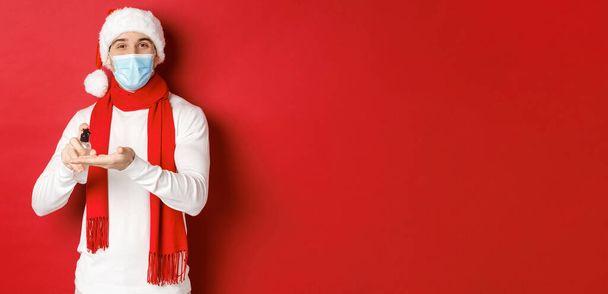 Концепция ковида-19, Рождества и праздников во время пандемии. Счастливый молодой человек в шляпе Санты и медицинской маске, дезинфицирует руки антисептиком и улыбается, стоя на красном фоне - Фото, изображение