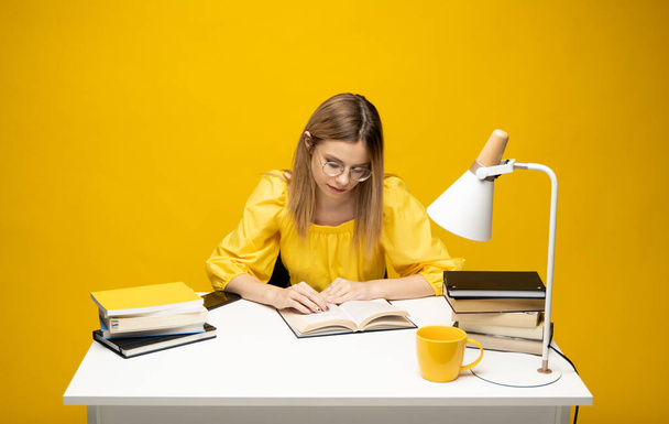 Young student woman in yellow casual clothes reading the book in library of university or college. Sitting and reading on yellow background. Studying. - 写真・画像