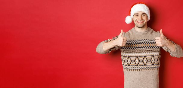 Изображение веселого привлекательного мужчины в зимнем свитере и шляпе Санты, показывающего большие пальцы вверх, празднующего Рождество и выглядящего счастливым, как нечто, стоящего на красном фоне - Фото, изображение