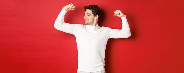 Πορτρέτο του χαμογελαστού όμορφος άντρας σε λευκό πουλόβερ, κάμψη δικέφαλους και κομπορρημοσύνη με δύναμη, επίδειξη ισχυρή μυς μετά την προπόνηση, στέκεται πάνω από το κόκκινο φόντο - Φωτογραφία, εικόνα