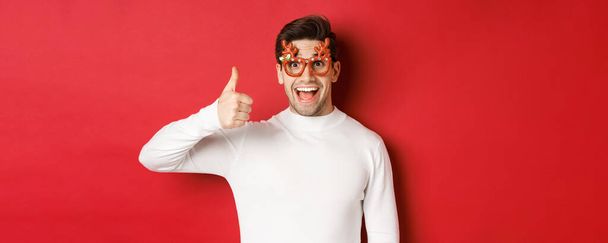 Zbliżenie przystojnego młodzieńca w okularach imprezowych i białym swetrze, z kciukiem do góry, cieszącego się wakacjami noworocznymi, stojącego nad czerwonym tłem - Zdjęcie, obraz