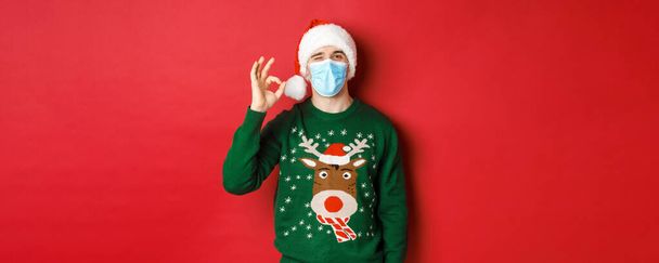 Koncept nového roku, covid-19 a sociální distancování. Veselý muž v lékařské masce a Santa klobouku, ukazující dobré znamení, doporučující něco dobrého, stojící nad červeným pozadím - Fotografie, Obrázek