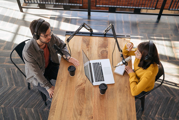 Zwei junge Radiomoderatoren mit Kopfhörern unterhalten sich, während sie neben Mikrofonen sitzen und im Sendestudio einen Audio-Podcast machen. Ansicht von oben. Soziale Medien, Podcast-Erstellung, Content Creator, Blogging - Foto, Bild
