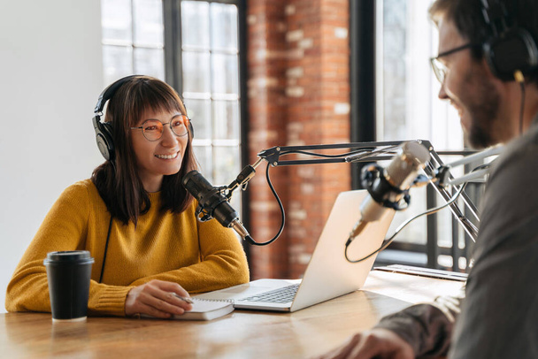 Young beautiful woman radio hosts smiling while interviewing man guest in studio while recording podcast for online show. Two podcasters in headphones looking at each other and recording audio podcast - Fotoğraf, Görsel