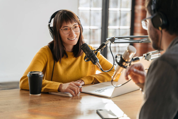 Podcasting. Man and woman podcasters interview each other for radio podcast in studio. Pretty female host in headphones smiling and looking on male guest while interviewing him for online show - Photo, Image