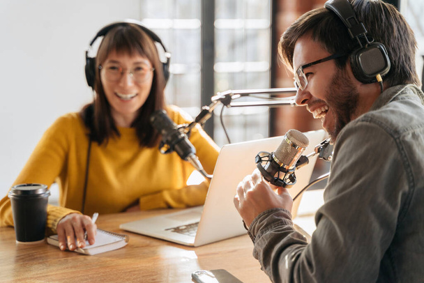 Selective focus of cheerful radio host laughing while recording podcast with colleague. Two radio hosts laughing while sitting near microphones in broadcasting studio and making audio podcast - 写真・画像