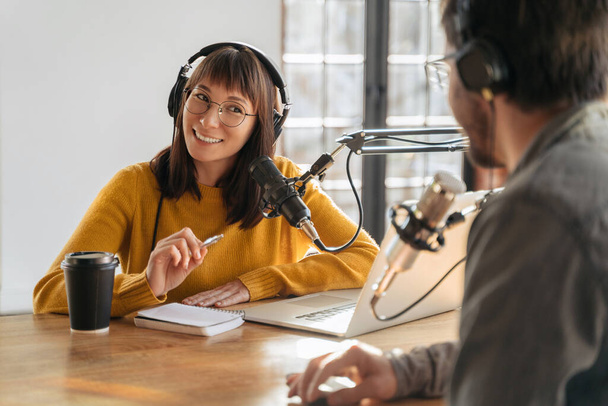 Radio hosts in headphones recording podcast in studio together. Cheerful woman host looking at man guest, smiling, holding a pen, interviewing an influencer. Podcasting and blogging, internet job - Foto, immagini