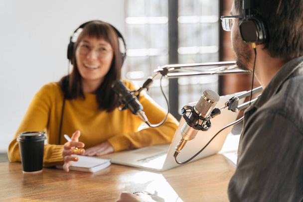 Two young podcasters in headphones recording and broadcasting podcast in studio. Woman and man making live audio podcast, running their radio show using microphones. Selective focus on microphone - 写真・画像