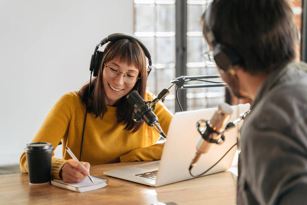 Vrouwelijke podcaster in hoofdtelefoon interviewt man gast met behulp van microfoon en laptop, schrijven in notebook, glimlachen, opnemen en uitzenden audio podcast in de studio. Inhoudmaker, bloggen - Foto, afbeelding