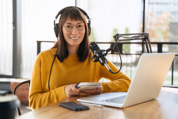 Πορτρέτο της χαρούμενης γυναίκας υποδοχής streaming audio podcast χρησιμοποιώντας μικρόφωνο και laptop στο στούντιο εκπομπής, κρατώντας σημειωματάριο, κοιτάζοντας κάμερα και χαμογελώντας. Νεαρή γυναίκα podcaster τρέχει ραδιοφωνική εκπομπή της - Φωτογραφία, εικόνα