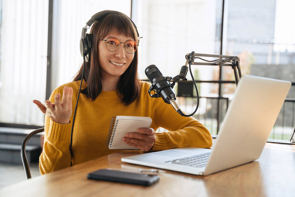 Gyönyörű női podcaster fejhallgatóval és szemüveggel gesztikulál, miközben audio podcast-ot sugároz a stúdióban, interjút készít egy mellénnyel, felé néz. Fiatal nő blogger beszél egy mikrofonba - Fotó, kép