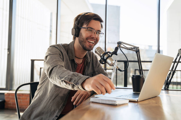 Νεαρός οικοδεσπότης σε ακουστικά και γυαλιά απολαμβάνοντας podcasting στο στούντιο, μιλώντας σε ένα μικρόφωνο, κρατώντας ένα στυλό, χρησιμοποιώντας φορητό υπολογιστή. Όμορφος podcaster γελώντας ενώ streaming ζωντανή μετάδοση ήχου podcast - Φωτογραφία, εικόνα