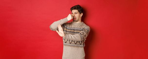 Imagen de hombre guapo disgustado en suéter de invierno, rechazando algo con mal olor, cerrar la nariz y levantar la mano para declinar, de pie sobre el fondo rojo - Foto, imagen