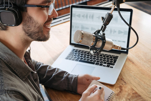 Cheerful host streaming his audio podcast using microphone and laptop, holding a pen and notepad. Close-up. Smiling podcaster in headphones and glasses enjoying making podcast, interviewing a gues - Foto, Imagem
