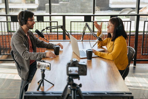 Two young bloggers, man and woman in headphones and glasses looking at each other while talking, recording conversation, interview for video blog. Female host interviewing guest for her video podcast - Photo, Image