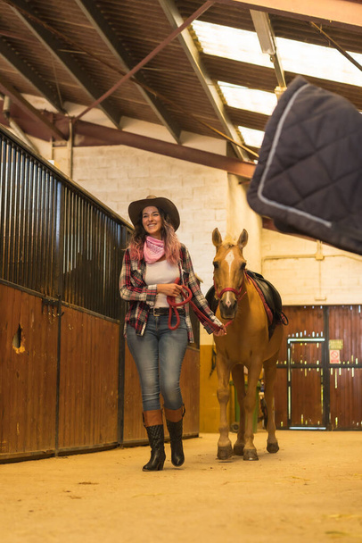 Caucasian cowgirl woman posing and smiling with a horse in a stable, southern usa hats, pink plaid shirt and jeans - 写真・画像