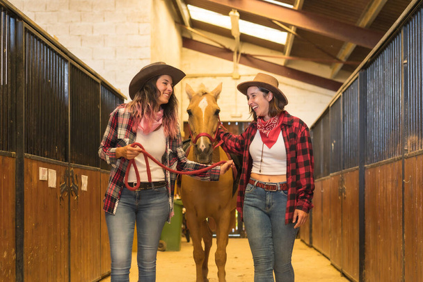 Two cowgirl women having fun and laughing with a horse in a stable, with South American outfits - Valokuva, kuva