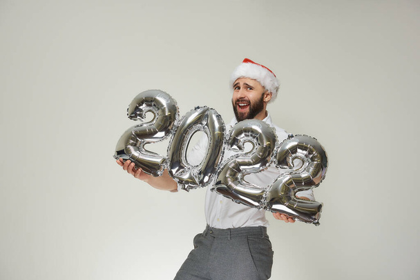 A dashing man in a red velvet Santa hat is holding silver balloons in the shape of 2022. A smiling guy with a beard at a New year party. - Photo, Image