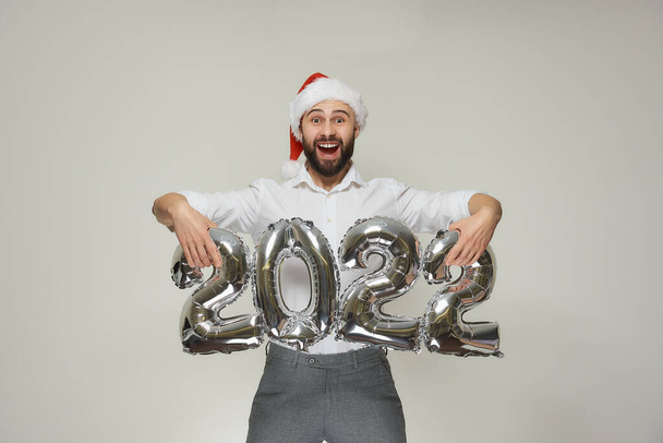 Een gelukkige man in een rood fluwelen Kerstmuts tilt zilveren ballonnen op in de vorm van 2022. Een blije kerel met een baard op een nieuwjaarsfeest. - Foto, afbeelding